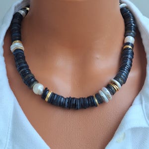 Könnte beinhalten: Eine dunkelgraue Perlenkette mit weißen und goldenen Akzenten. Die Halskette besteht aus dunkelgrauen, scheibenförmigen Perlen, die mit weißen Perlen und goldfarbenen Abstandshaltern durchsetzt sind. Die Halskette wird auf einer Schaufensterpuppe präsentiert.