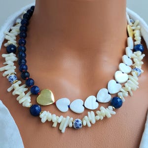 Op de afbeelding: Een gelaagde ketting met witte koraalachtige elementen, marineblauwe kralen en hartvormige witte schelpaccenten. Gouden hartvormige bedels en blauw-witte porseleinen kralen voegen details toe aan deze sieraden.