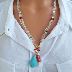 May include: A gold-toned necklace with clear, rectangular beads and accents of turquoise and red stones. The necklace features a teardrop-shaped turquoise pendant and a red, cylindrical bead pendant. The necklace is worn over a white collared shirt.