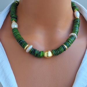 Op de afbeelding: Een ketting bestaande uit een streng kleine, platte, ronde groene kralen afgewisseld met onregelmatige witte parels en goudkleurige tussenstukjes. Een enkele, grotere, gladde, goudkleurige bolvormige kraal bevindt zich in het midden.