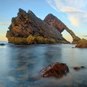 Puede incluir: Una gran formación rocosa con un arco natural, creando un marco para el cielo azul y las nubes. La roca es una mezcla de marrón, naranja y verde, con el agua en primer plano de un azul suave.