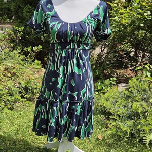 Puede incluir: Vestido azul marino con estampado floral verde y blanco, exhibido en un maniqu&iacute; blanco. El vestido tiene mangas cortas, escote redondo y falda con volantes. Fabricado con tejido ligero, es ideal para climas c&aacute;lidos.