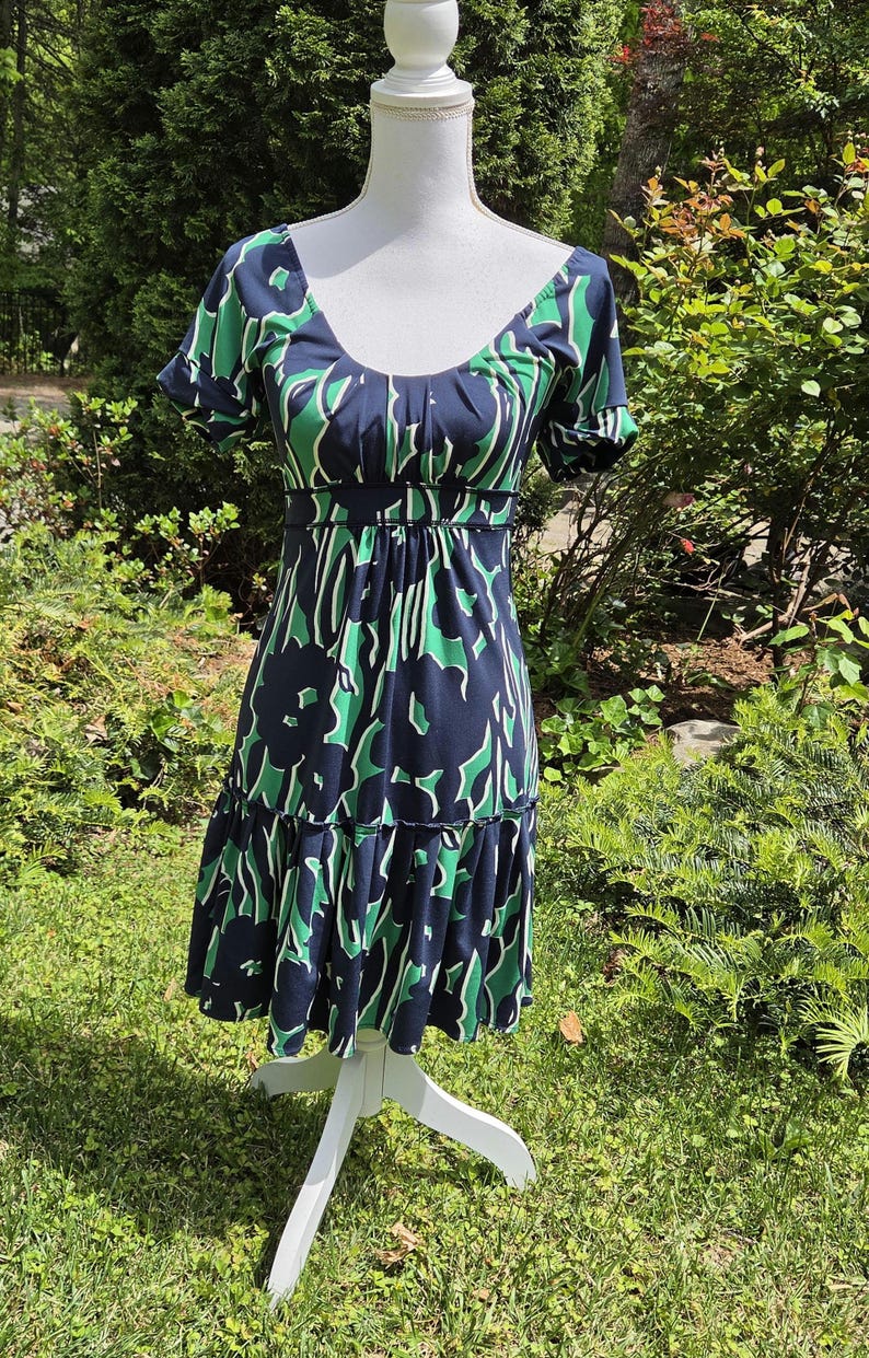 Puede incluir: Vestido azul marino con estampado floral verde y blanco, exhibido en un maniqu&iacute; blanco. El vestido tiene mangas cortas, escote redondo y falda con volantes. Fabricado con tejido ligero, es ideal para climas c&aacute;lidos.