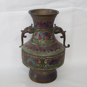 May include: A decorative brass vase with a floral design in shades of blue, red, and green. The vase has two handles shaped like elephant heads.