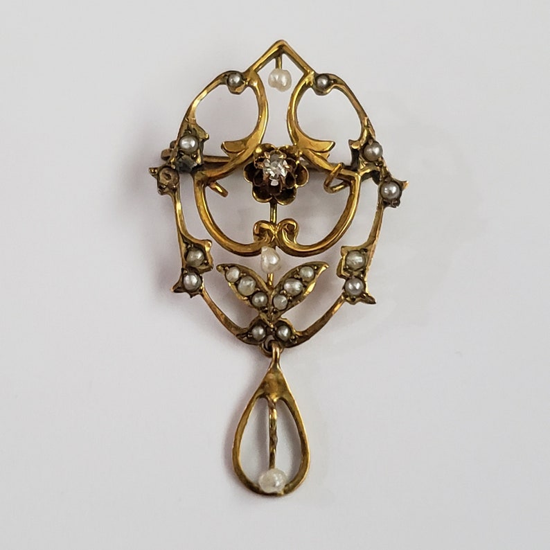 May include: A gold pendant brooch with a teardrop-shaped design. The pendant is adorned with a small diamond and pearl accents. The brooch is in the shape of a stylized flower.
