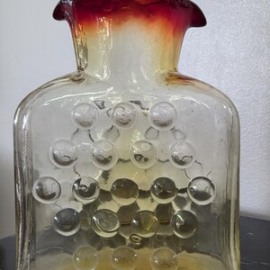 May include: A clear glass vase with a square body and a flared, ruffled rim. The rim transitions from red to yellow. The body features a pattern of raised, clear glass circles. The vase is likely for decorative purposes.
