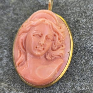 May include: Oval pendant with a carved cameo of a woman's profile in a light coral color. The cameo is set in a gold-colored frame with a small loop at the top for a chain. The background is a textured gray surface.