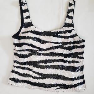 May include: A sequined tank top with a zebra stripe pattern in black and silver. The top has a scoop neckline and wide straps. The fabric is covered in small, reflective sequins, creating a shimmering effect. The garment is likely for special occasions.