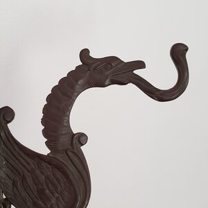 May include: A dark brown metal dragon-shaped hook with a curled tail and a detailed head with a long beak. The hook is designed to be mounted on a wall.