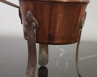 May include: A small copper-colored pot with a handle and three metal legs. The pot has a shiny finish and decorative metal accents. The legs are attached to the pot with rivets. The pot is designed for cooking or serving.