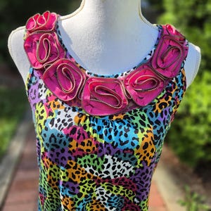 May include: A sleeveless top featuring a vibrant, multi-colored animal print pattern in shades of blue, purple, yellow, and green. The round neckline is embellished with a series of bright pink fabric rosettes constructed from coiled zippers.