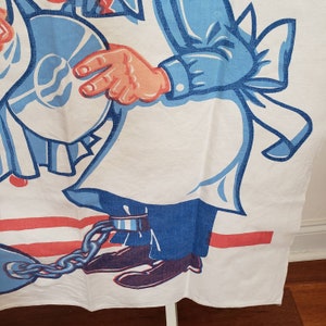 May include: A white cotton tea towel with a cartoon illustration of two people wearing blue and white clothing. The person on the right is holding a blue plate and is chained to a metal ball and chain. The background is red and white stripes.