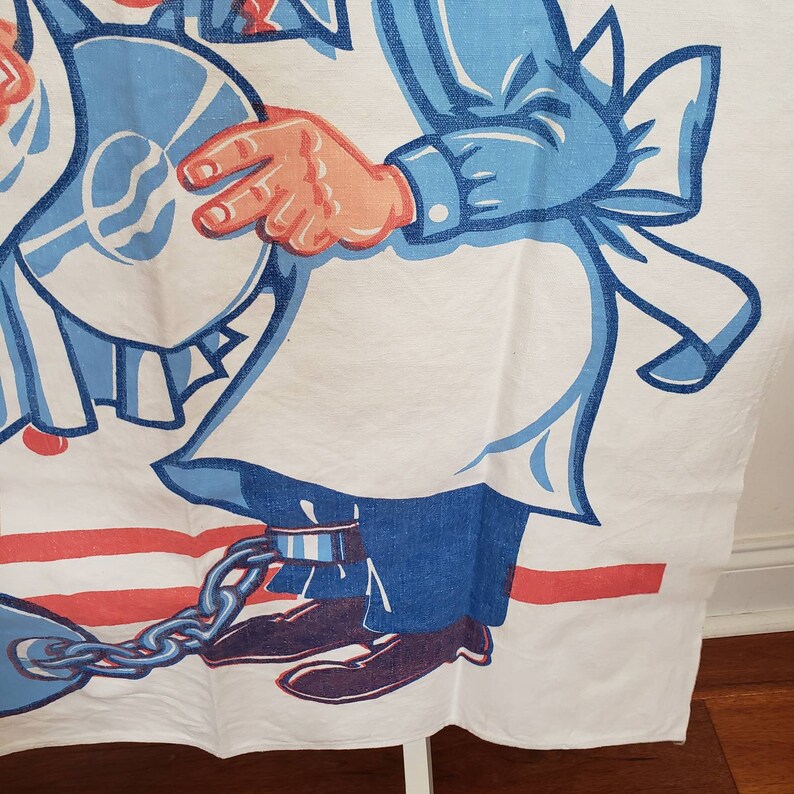 May include: A white cotton tea towel with a cartoon illustration of two people wearing blue and white clothing. The person on the right is holding a blue plate and is chained to a metal ball and chain. The background is red and white stripes.