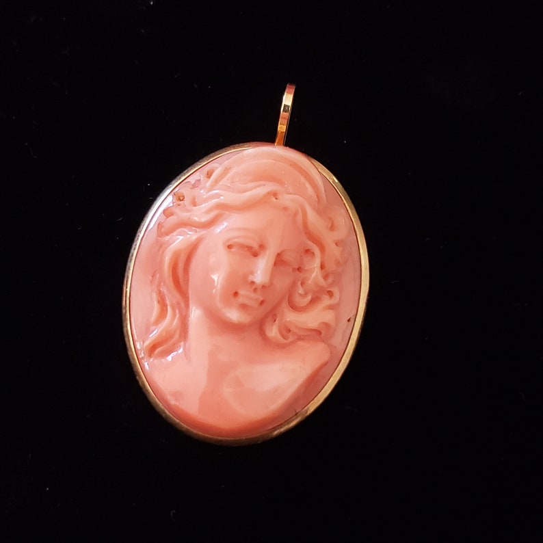 May include: Oval pendant with a carved coral cameo depicting a woman's profile. The cameo is set in a gold-colored frame and has a loop at the top for a chain. The coral has a peach hue, and the background is black.