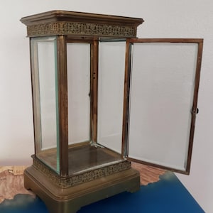 K&ouml;nnte beinhalten: Eine Vitrine aus Messing mit einer offenen Glast&uuml;r. Die Vitrine hat einen dekorativen oberen und unteren Rand. Die Vitrine steht auf einer blauen Oberfl&auml;che.