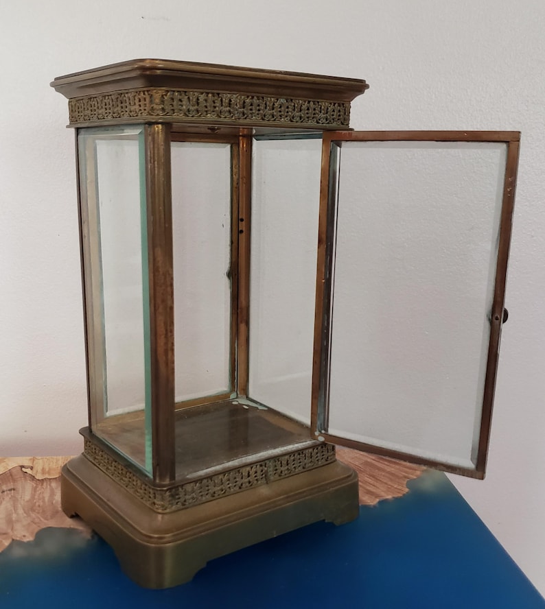 K&ouml;nnte beinhalten: Eine Vitrine aus Messing mit einer offenen Glast&uuml;r. Die Vitrine hat einen dekorativen oberen und unteren Rand. Die Vitrine steht auf einer blauen Oberfl&auml;che.