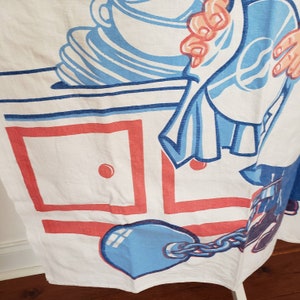 May include: A white kitchen towel with a blue and red illustration of a person holding a dish towel and a blue ball and chain. The illustration is on a white background with red and blue squares.