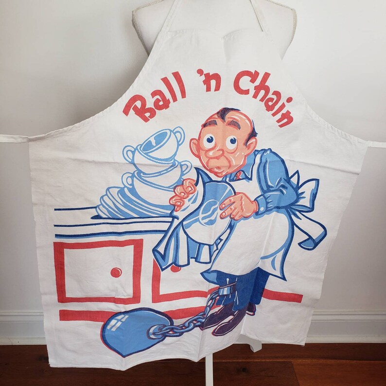 May include: White kitchen apron with a cartoon illustration of a man in a blue shirt and white pants. The man is holding a plate of dishes and is chained to a ball. The apron has the text "Ball 'n Chain" in red and blue letters.