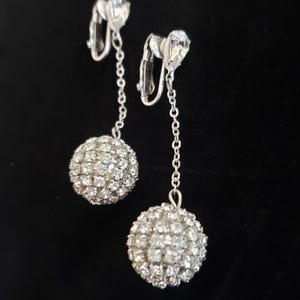 May include: A pair of silver clip-on earrings with dangling crystal ball charms. The balls are covered in small, clear rhinestones.
