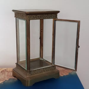 K&ouml;nnte beinhalten: Eine Vitrine aus Messing mit einer Glast&uuml;r und einer Glasfront. Die Vitrine hat einen dekorativen Rahmen um die Ober- und Unterseite.