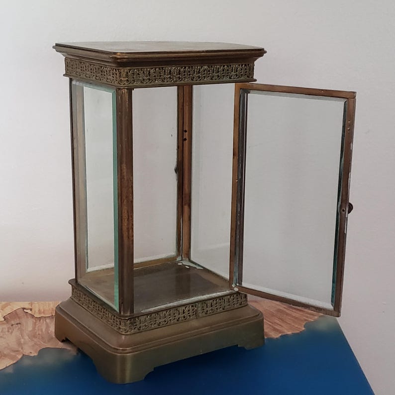 K&ouml;nnte beinhalten: Eine Vitrine aus Messing mit einer Glast&uuml;r und einer Glasfront. Die Vitrine hat einen dekorativen Rahmen um die Ober- und Unterseite.