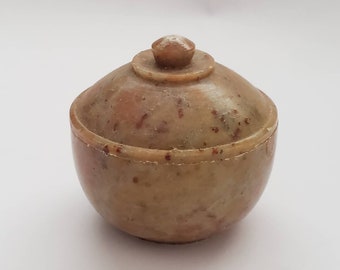 May include: A small, round, lidded container made of light brown stone with red speckles. The lid has a small knob on top.