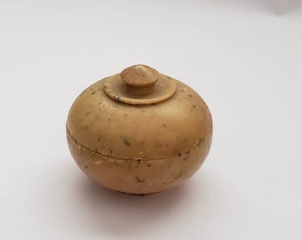 May include: A small, round, beige-colored container with a lid. The container is made of a material that looks like bone or ivory and has a speckled texture.