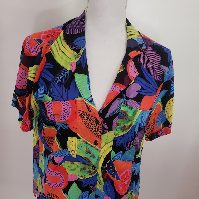 May include: A short-sleeved button-up shirt with a colorful abstract print featuring a variety of fruits, leaves, and geometric shapes. The shirt is made of a lightweight fabric and has a relaxed fit.