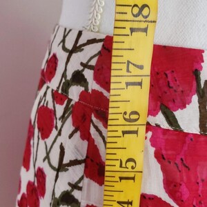 May include: A close-up of a white skirt with a red floral pattern and brown stems. A yellow measuring tape is placed vertically along the skirt, showing measurements in inches. The skirt is made of a lightweight fabric.