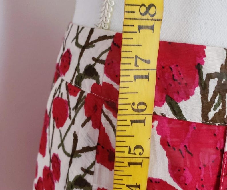 May include: A close-up of a white skirt with a red floral pattern and brown stems. A yellow measuring tape is placed vertically along the skirt, showing measurements in inches. The skirt is made of a lightweight fabric.