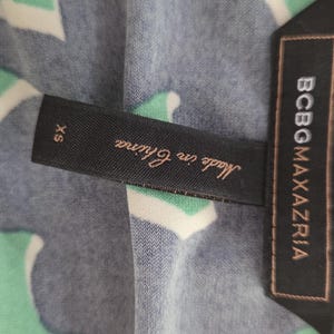 Puede incluir: Primer plano de una tela estampada con una etiqueta que dice "BCBGMAXAZRIA" y "Made in China". La tela tiene un dise&ntilde;o de formas blancas, verdes y azul marino. La etiqueta tambi&eacute;n indica la talla XS.