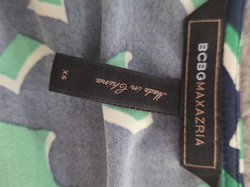 Puede incluir: Primer plano de una tela estampada con una etiqueta que dice "BCBGMAXAZRIA" y "Made in China". La tela tiene un dise&ntilde;o de formas blancas, verdes y azul marino. La etiqueta tambi&eacute;n indica la talla XS.