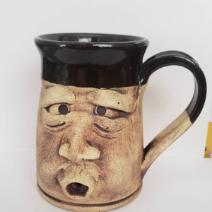 May include: A handmade ceramic mug with a sculpted face design. The mug features a brown rim and handle, with a tan body and a detailed, expressive face. The face has carved eyes, a nose, and an open mouth.
