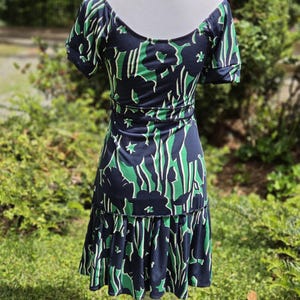 Puede incluir: Vestido azul marino con estampado floral verde y blanco. El vestido tiene mangas cortas, cuello redondo y cintura entallada. La falda tiene un dobladillo con volantes. El vestido se exhibe en un maniqu&iacute; blanco.