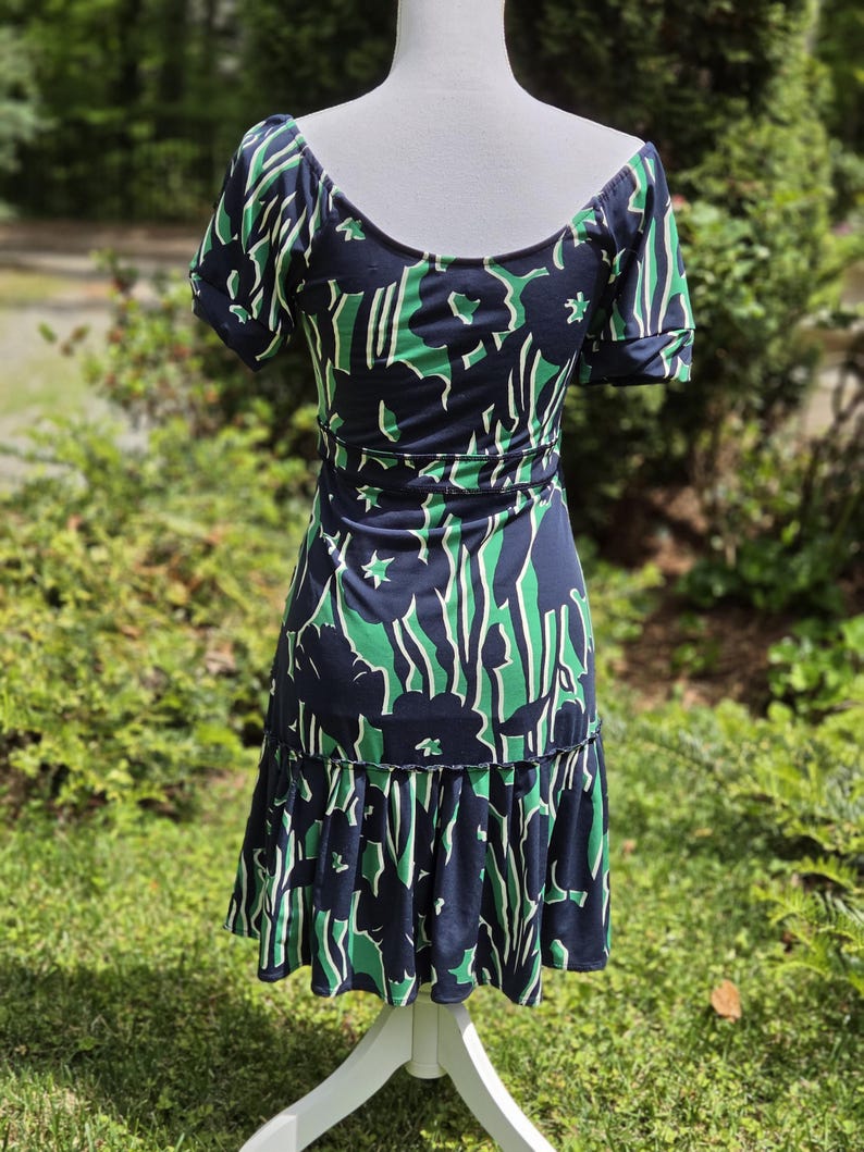 Puede incluir: Vestido azul marino con estampado floral verde y blanco. El vestido tiene mangas cortas, cuello redondo y cintura entallada. La falda tiene un dobladillo con volantes. El vestido se exhibe en un maniqu&iacute; blanco.