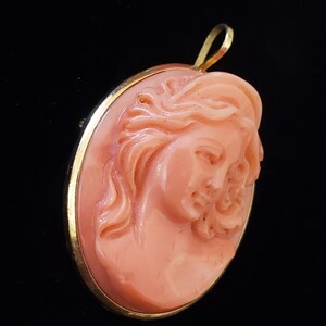 May include: Oval pendant featuring a carved cameo of a woman's profile in a peach hue. The cameo is set in a gold-colored frame with a loop at the top for a chain. The background is black.