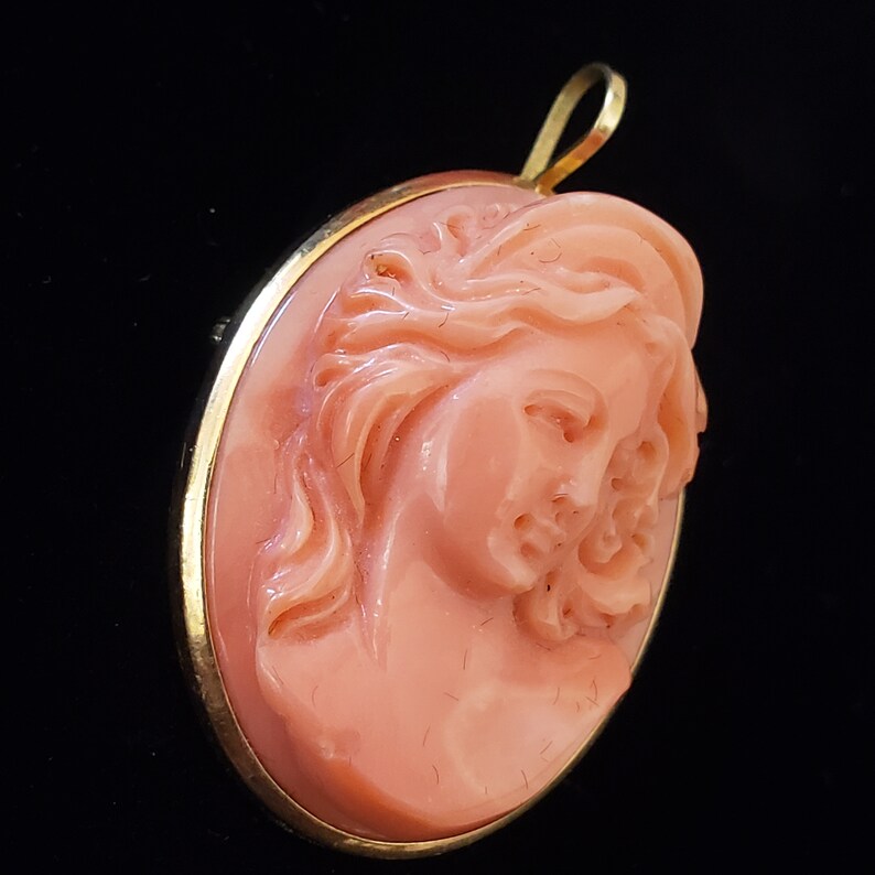 May include: Oval pendant featuring a carved cameo of a woman's profile in a peach hue. The cameo is set in a gold-colored frame with a loop at the top for a chain. The background is black.