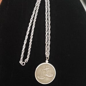 May include: A silver chain necklace with a round pendant featuring a silver Libra zodiac symbol. The pendant is engraved with the word "Libra".