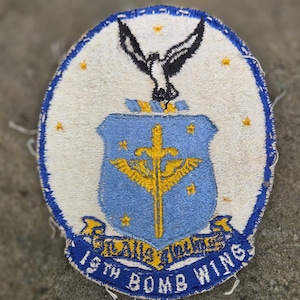 May include: An oval embroidered patch featuring a white background with a blue border. It displays a black bird above a light blue shield containing a yellow sword and wings. Below the shield is a blue banner with the text 19TH BOMB WING.