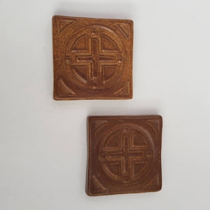May include: Two square ceramic tiles with a brown glaze. Each tile features a raised design of a circle with a cross inside. The cross is made up of four smaller circles.