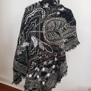 May include: A black shawl featuring intricate embroidery in shades of gray, white, and muted red. The fabric displays a complex pattern of leaves, geometric shapes, and swirling lines, finished with a frayed edge along the bottom hem.