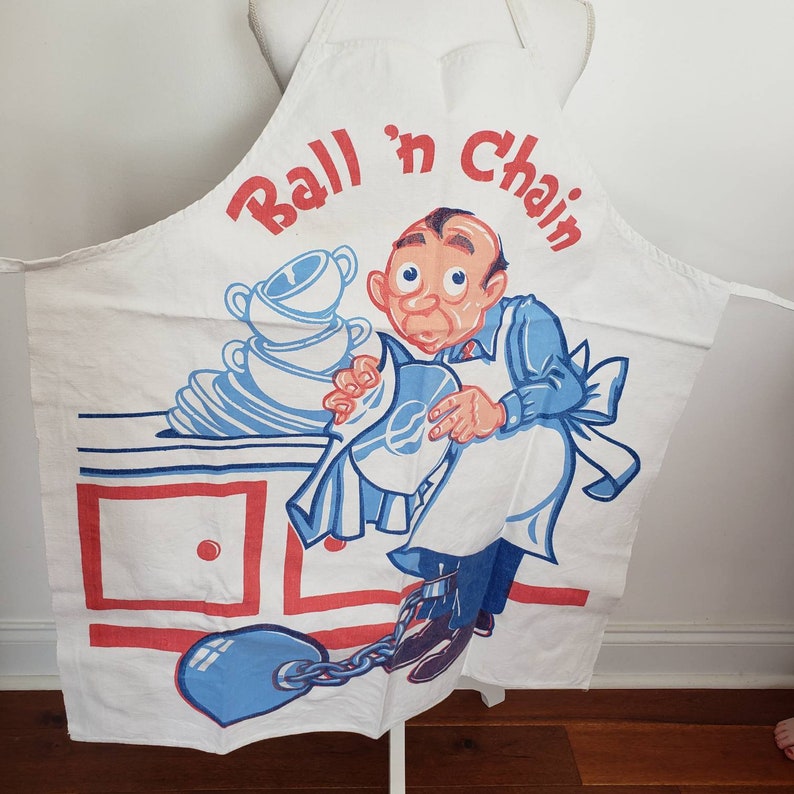 May include: A white apron with a cartoon illustration of a man in a blue shirt and white pants holding a blue ball and chain. The apron has the text "Ball 'n Chain" in red and blue letters. The man is standing in front of a white cabinet with two drawers. The apron has a blue ball and chain attached to the bottom.