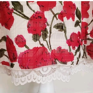 May include: A white fabric with a red floral pattern and green stems. The fabric has a delicate white lace trim along the bottom edge. The floral design features large, vibrant red flowers, creating a striking contrast against the white background.
