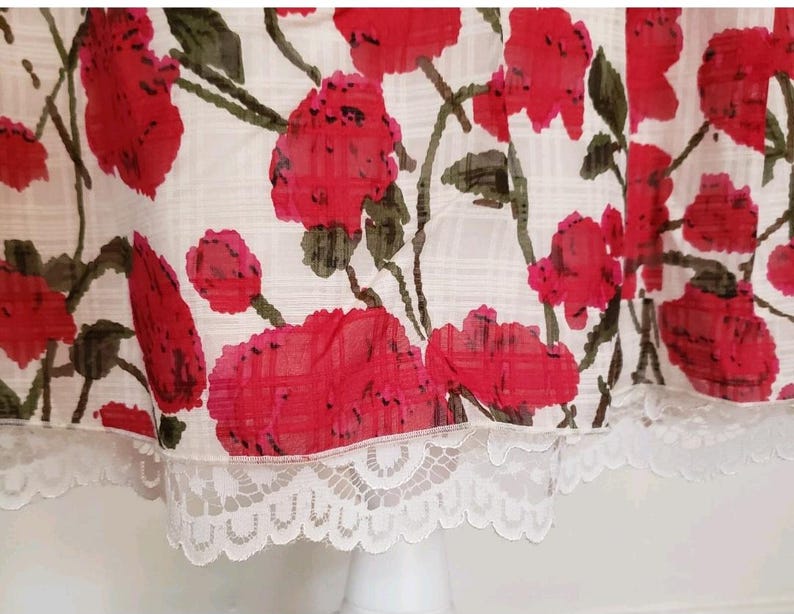 May include: A white fabric with a red floral pattern and green stems. The fabric has a delicate white lace trim along the bottom edge. The floral design features large, vibrant red flowers, creating a striking contrast against the white background.
