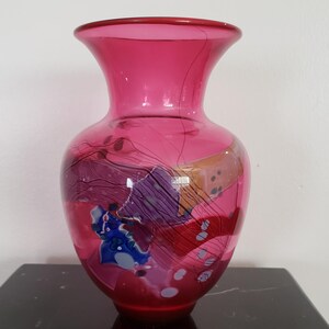 May include: A vibrant, hand-blown glass vase in shades of pink and red. The vase features abstract, colorful designs in blue, purple, and gold. The vase has a wide body and a flared top, perfect for displaying flowers.