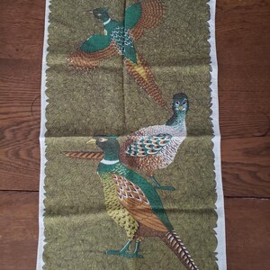 May include: A white tea towel with a green background and a brown border. The tea towel features a design of two pheasants with brown, green, and yellow feathers.