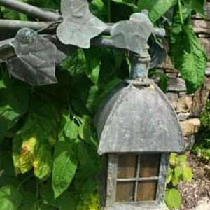 May include: Weathered metal outdoor lantern with a small window and a peaked roof. The lantern is attached to a metal rod with decorative leaf accents. The background features lush green foliage and a stone wall.