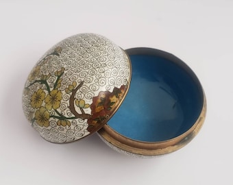 May include: A small, round, white and blue enamel trinket box with a floral design. The lid is open and the inside of the box is blue.