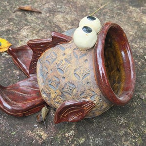 May include: A ceramic fish-shaped planter with a glossy dark brown rim and fins. The body features a textured, tan-colored geometric pattern and two protruding, round, off-white eyes with black pupils positioned on the top of the head.