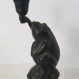 May include: Black cast iron frog candle holder. The frog is sitting on a base and holding a candle cup in its right hand.